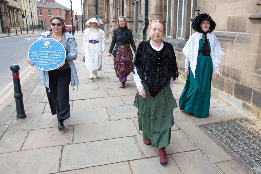 Women in Victorian costume waling down a road in Wakefield with Ann Hurst's Blue Plaque