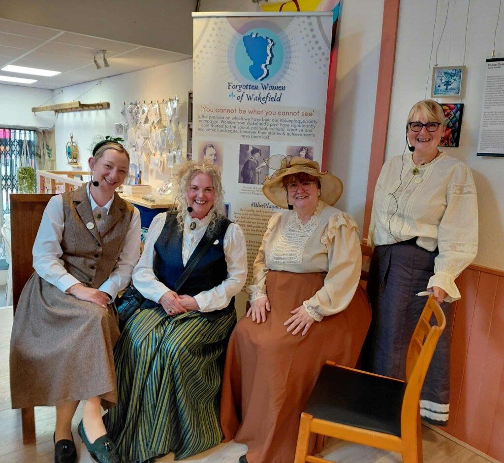 Women in Victorian dress smile at the camera at a commemoration event. 