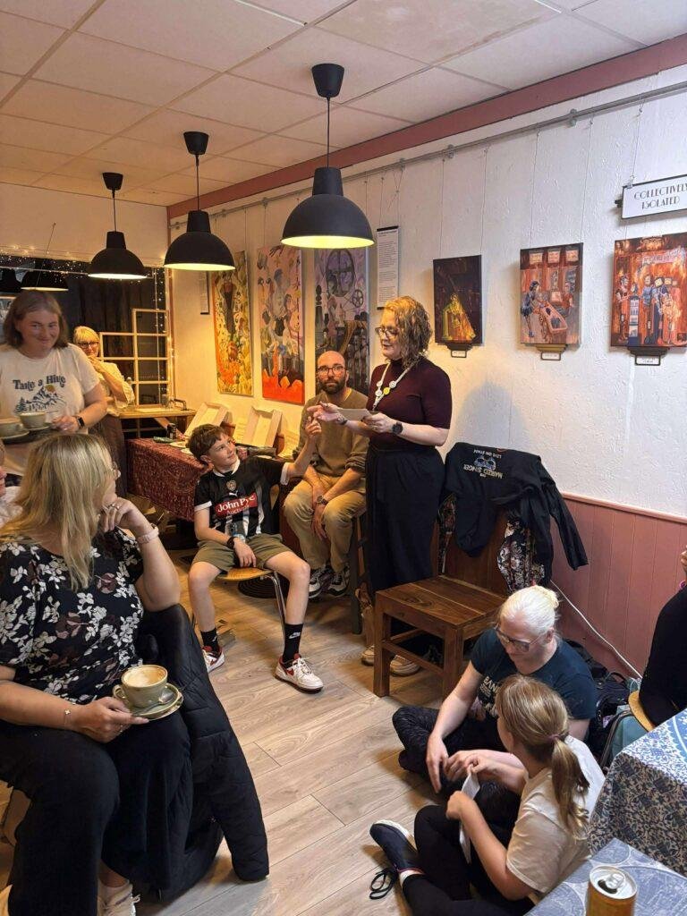 A cozy and yet somehow industrial looking cafe in which a lady is stood giving a talk to people. 