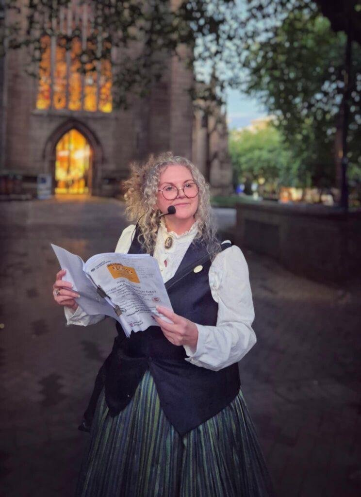 A smiling woman with glasses and curly hair is dressed in Victorian clothes. She is holding a pamphlet and has a radio mic. Behind her is the lit doors and windows of Wakefield Cathedral. 