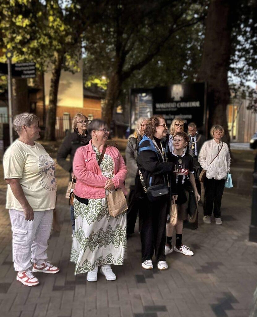 A group of people stand listening to a talk by someone in Victorian dress. 