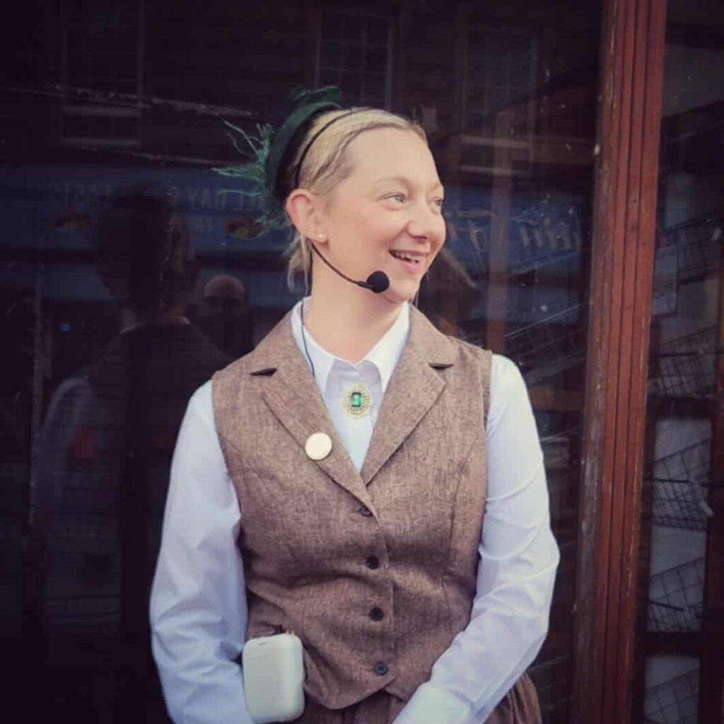 A smiling woman in Victorian waistcoat and dress gives a talk on a radio mic. 